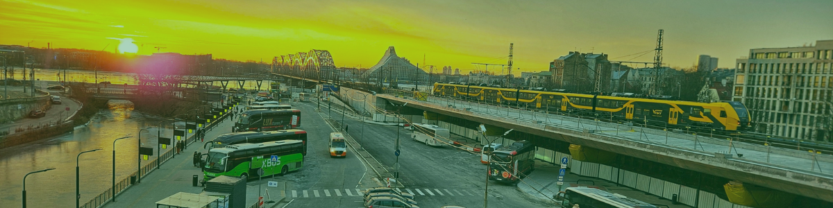 Attēlā redzams panorāmas skats no augšas uz Rīgas starptautiskās autoostas apkārtni, kur apakšā stāv rindā vairāki starppilsētu un tūrisma autobusi. Pa labi, augstākajā līmenī, atrodas dzelzceļa sliedes ar garu, dzeltenu vilcienu, kas šķērso pilsētas centru. Kreisajā malā redzama upes mala ar piestātnēm, bet fonā slejas vēsturiskais metāla dzelzceļa tilts un citi Rīgas ēku silueti. Priekšplānā redzama krustojuma daļa un ietves, kur cilvēki pārvietojas starp autoostu un apkārtējiem objektiem. Gaisā ir izteikts dzeltenīgs saulrieta tonis, apgaismojot visu panorāmu siltās nokrāsās.