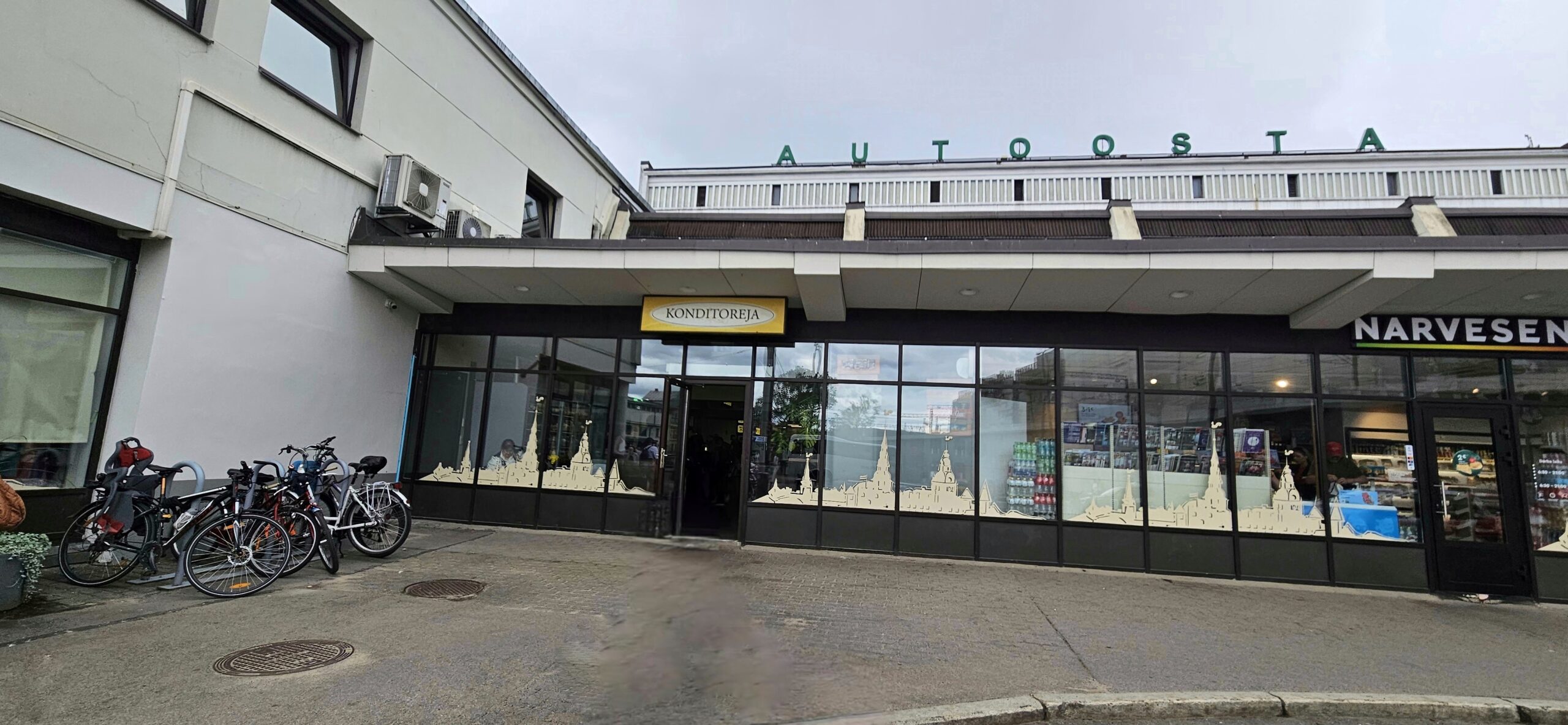 Attēlā redzams ēkas fasādes skats ar gaišu cokolu un jumta daļu, uz kuras lieliem, zaļiem burtiem ir uzraksts “AUTOOSTA”. Zem jumta pārkares ir vairāki iestāžu logi un durvis. Centrā atrodas ieeja ar dzeltenu izkārtni “KONDITOREJA”, uz logiem uzlīmēts pilsētas panorāmas siluets. Labajā malā redzama “Narvesen” veikala fasāde ar stiklotu ieeju. Kreisajā malā pie ēkas novietoti vairāki divriteņi. Ielas segums ir asfaltēts, neliels stūra nobraukums norāda uz auto piebraucamo ceļu. Kopumā fotogrāfijā redzama Rīgas starptautiskās autoostas ēkas ārtelpa ar konditorejas ieeju, velonovietni un blakus esošu “Narvesen” stendu.