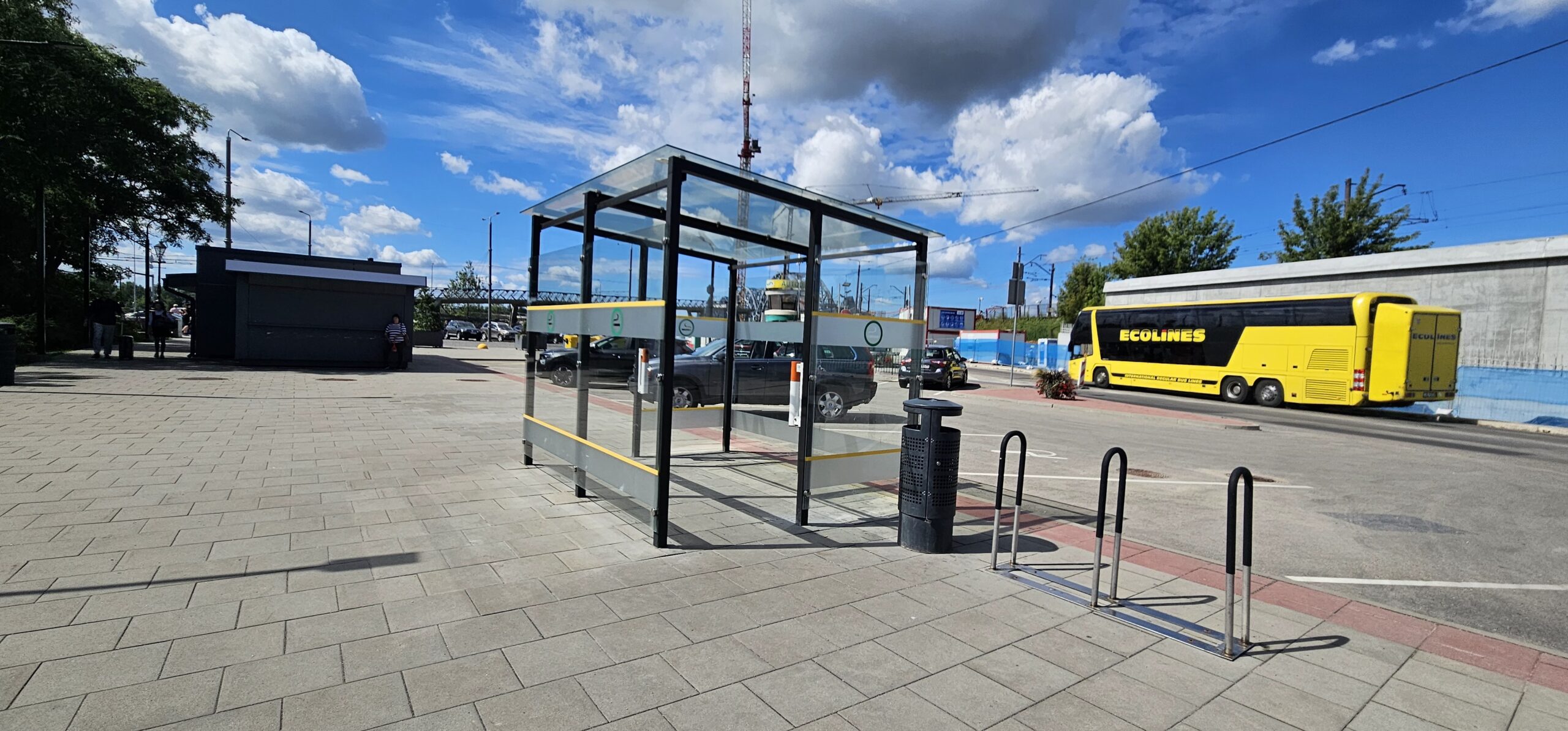 Attēlā redzama neliela, stiklotas konstrukcijas autobusa pietura pilsētas vidē. Tā atrodas uz platas, gaišas flīžu vai betona seguma ietves. Pieturas sienas ir no caurspīdīga stikla, nostiprinātas metāla rāmī, un tai apkārt izvietotas dzeltenas un melnas drošības svītras. Blakus pieturai redzams tumšs atkritumu tvertne un metāla statīvs velosipēdu pieslēgšanai.  Attēla fonā, labajā pusē, stāv koši dzeltens starppilsētu autobuss ar uzrakstu “Ecolines”. Nedaudz tālāk redzama siena ar zilu nokrāsu, kā arī pilsētas ainava ar dažām ēkām un mastu, kas iespējams, ir domāts televizora vai radio raidstacijas torņa vajadzībām. Debesīs ir lieli, balti mākoņi, un kopumā laiks šķiet saulains, spriežot pēc zilās debess un gaišās, spilgtās gaismas. Pa kreisi no pieturas redzama tumša, neliela ēka vai kiosks, bet kreisajā malā daļēji var saskatīt arī kokus.