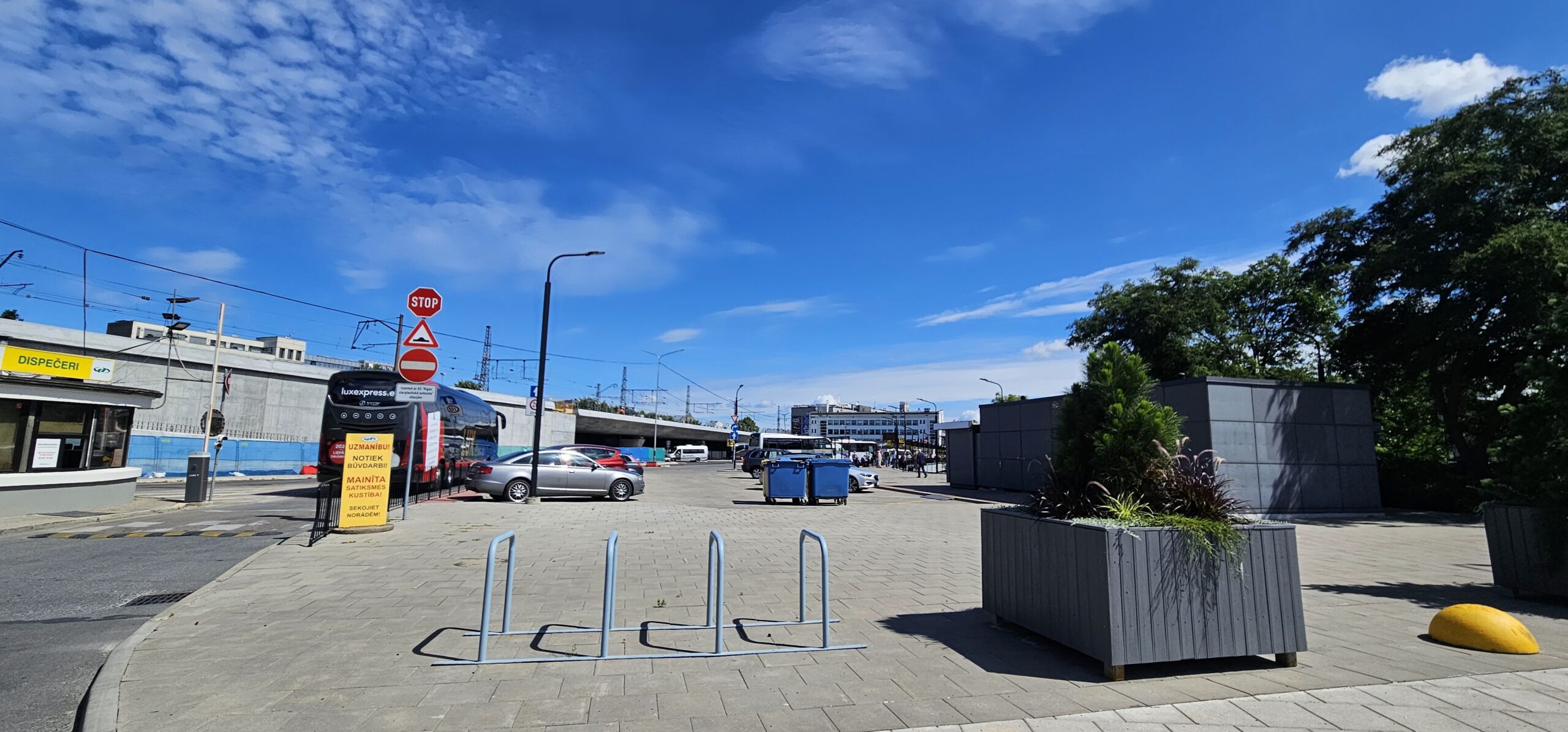Attēlā redzams plašs, gaiši bruģēts laukums pie Rīgas starptautiskās autoostas, skats vērsts virzienā, kur labajā pusē slejas nelieli pelēki būvmateriālu blokveida kioski vai noliktavas tipa ēkas. Pa vidu, tuvāk priekšplānam, atrodas metāla velostatīvs un liels koka puķu kaste ar zaļiem augiem. Kreisajā pusē redzams augsts metāla stabs ar vairākām ceļa zīmēm, tostarp sarkanu “STOP” un “Iebraukt aizliegts” norādēm, kā arī turpat novietota liela, dzeltena informācijas plāksne. Aiz tās zilganpelēkā tonī atrodas zema betona siena (iespējams, nožogojums), aiz kuras redzams dzelzceļa uzbērums un stacijas infrastruktūra ar elektriskajiem kontaktiem un sliežu ceļiem.  Tālāk fonā paveras Rīgas starptautiskās autoostas ēkas un to peroni, kur redzami arī zilganbalti vagoni vai autobusi. Debesis ir gaišas, dzidri zilas, ar lieliem, baltiem mākoņiem. Labajā malā, netālu no pelēkā kioska, redzami daļēji apstādījumi un koku lapotne, kas papildina pilsētvides zaļumu. Vispārējais iespaids ir par plašu, sakārtotu zonu, kas paredzēta gājēju un transporta kustībai starp autoostu un tās apkārtni.