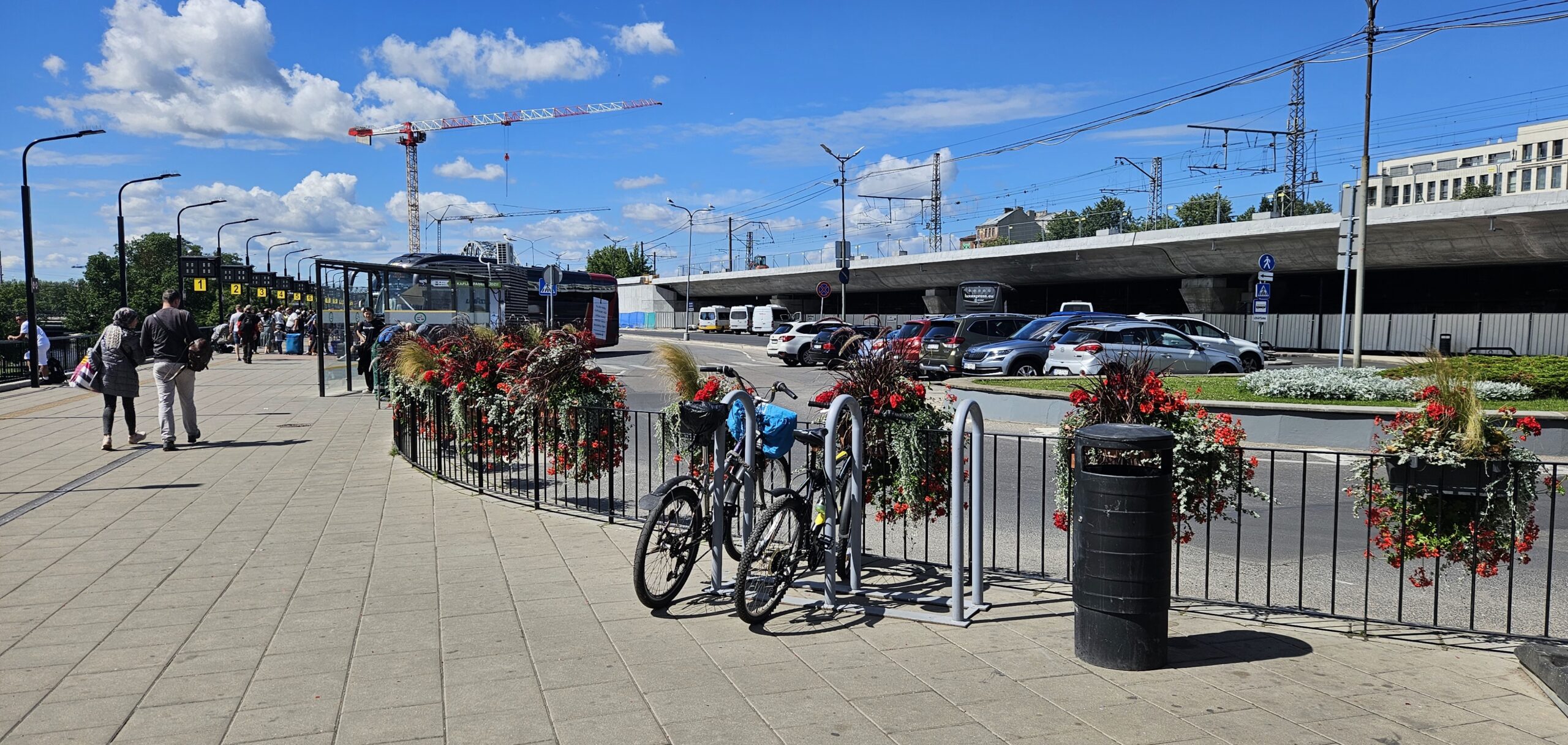 Atrašanās vieta: Plaša, bruģēta ietve (perons) pie autoostas.  Priekšplānā (ietves malā) vidū:  Velostatīvs (metāla rāmis), pie kura pieslēgti 2 velosipēdi.  Blakus velostatīvam melnas krāsas cilindriska atkritumu urna.  Zems metāla sētiņas posms ar uzkārtiem puķu groziem norobežo ietvi no brauktuves.  Tālāk pa kreisi:  Cilvēki stāv rindā vai pārvietojas, turpinās autobusa pieturas (stiklotas nojumes).  Pa labi (aiz sētiņas):  Brauktuve un stāvvieta automašīnām.  Lielākais ceļa pārvads (betona konstrukcija) atrodas vēl tālāk pa labi.  Ja vēlaties doties uz autobusiem, tie parasti atrodas pa kreisi gar stiklotajām pieturām. Uzmanieties no puķu nožogojuma un velostatīva – tie var būt kā šķērslis gaitai gar ietves malu.
