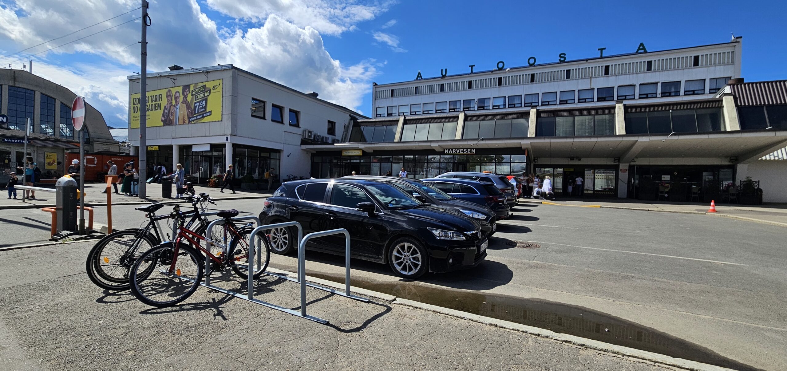 Atrašanās vieta: Foto redzama Rīgas starptautiskās autoostas galvenā ēka (plaša fasāde ar uzrakstu “AUTOOSTA” ēkas jumta malā).  Priekšplānā (autostāvvieta):  Starp braucamo daļu un ēku novietotas vairākas automašīnas.  Netālu no brauktuves malas atrodas velostatīvs, kur redzami vismaz 3 pieslēgti velosipēdi.  Ieeja autoostā:  Centrā ir galvenās durvis uz ēku (stiklotas ieejas), pa kreisi no durvīm pamanāms plašs stiklots laukums, pa labi – Narvesen kiosks.  Lai nonāktu līdz ieejai, jāpaiet gar automašīnām stāvvietā, tad jākāpj uz nelielas ietves/bruģētas platformas, kas ved pie ēkas durvīm.  Pa kreisi no ēkas:  Atrodas atsevišķa ēka ar lielu reklāmas plakātu (tas nav tiešs ieejas punkts).  Īsumā: piesargieties no autostāvvietas, sekojiet ietvei, ejiet centrā virzienā uz galveno durvju rindu, kur atrodas ieeja autoostā, blakus Narvesen kioskam.