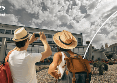 Divi cilvēki ar cepurēm un mugursomām stāv autoostas ārpusē, viens fotografē autoostas ēku - moderna daudzstāvu ēka ar stikla fasādi; debesis daļēji mākoņainas