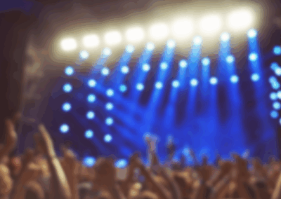 Koncerts ar spilgtu skatuves apgaismojumu un paceltām skatītāju rokām, zilā gaismā izgaismota skatuve un liels ekrāns fonā.