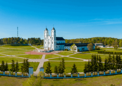 Panorāmas skats uz Aglonas baziliku Latvijā – balta divtorņu baznīca plašā zaļā teritorijā ar celiņiem, kokiem, žogu priekšplānā un mazākām ēkām fonā