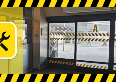 Slēgtas stikla durvis, A ieeja autoostā, ar brīdinājuma lentēm un uzlīmēm, kas norāda uz remontdarbiem, kreisajā pusē dzeltena ikona ar āmuru un atslēgu, fonā redzama iela caur stiklu