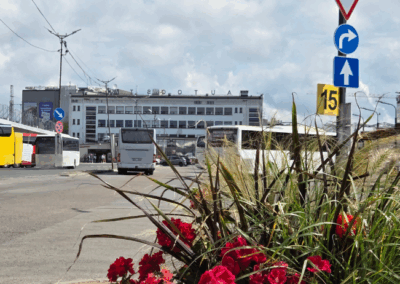 Rīgas starptautiskā autoosta ar stāvošiem autobusiem, redzama ceļa zīme ar pagriezienu pa labi un skaitli 15. Priekšplānā ir puķu pods ar sarkanām un zaļām puķēm, fonā – liela ēka un daļēji mākoņains debesjums.