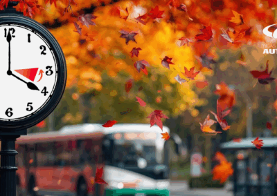 Melns pulkstenis ar baltiem cipariem rāda laiku 4:00, fonā rudens ainava ar krītošām sarkanām un oranžām lapām, autobusi un pietura, augšējā labajā stūrī redzams Rīgas Starptautiskās Autoostas logotips.