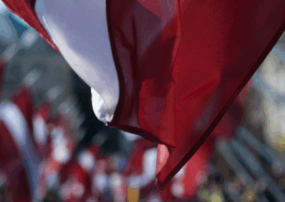 Latvijas karogs tuvplānā ar sarkanbaltsarkanu krāsu, fonā redzami vairāki citi Latvijas karogi, kas plīvo gaisā svinīgā pasākumā